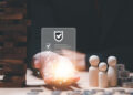 A hand holds a glowing shield icon with checkmarks above a table featuring stacked coins, wooden figures representing a family, and a Jenga tower, symbolizing financial security and protection. | LESSINVEST