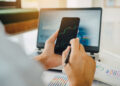 Person holding a smartphone displaying a stock chart, with a laptop and financial reports in the background. | LESSINVEST