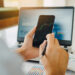 Person holding a smartphone displaying a stock chart, with a laptop and financial reports in the background. | LESSINVEST