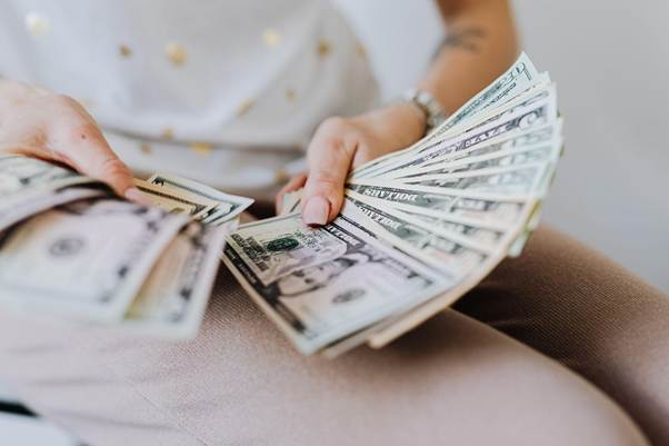 A person wearing light-colored pants and a white shirt is holding and counting a large stack of U.S. dollar bills, with various denominations visible. | LESSINVEST