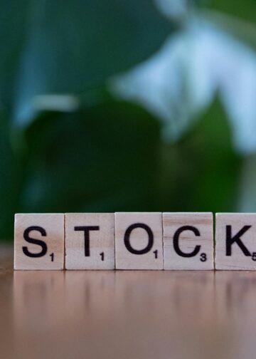 Wooden Scrabble tiles spell out the word "STOCK" on a smooth surface, with a blurred green leafy background. | LESSINVEST