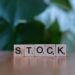Wooden Scrabble tiles spell out the word "STOCK" on a smooth surface, with a blurred green leafy background. | LESSINVEST