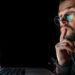 A man wearing glasses looks thoughtfully at a laptop screen displaying stock market charts. | LESSINVEST
